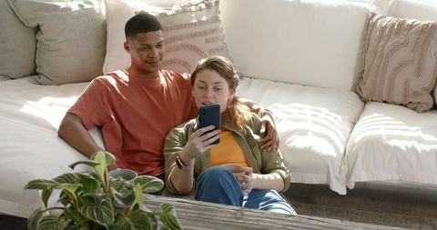 Couple cuddling on sunlit sectional sofa scrolling smartphone and enjoying cozy home