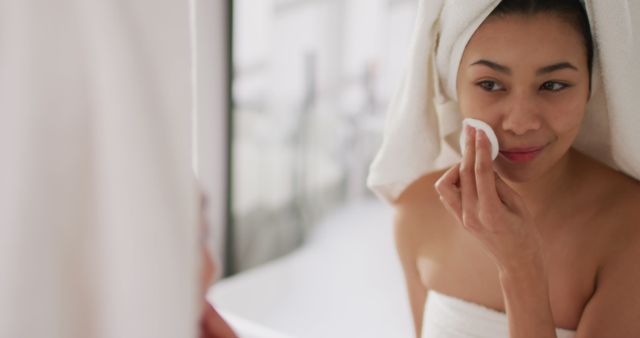 Image of portrait of smiling biracial woman with towel on hair cleansing face in bathroom. Health and beauty, leisure time, domestic life and lifestyle concept.
