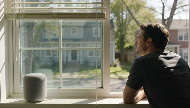 Leaning man gazing out window with smart speaker on sill in sunlit suburban home