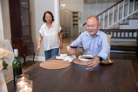 Senior asian couple sharing cozy wine dinner at home, man holding glass while woman walking
