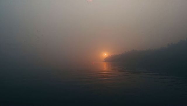 Misty dawn over calm water with orange sun reflection and silhouetted wooded headland