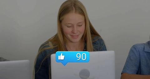 Smiling teen girl using laptop in classroom showing social thumbs-up 90 overlay