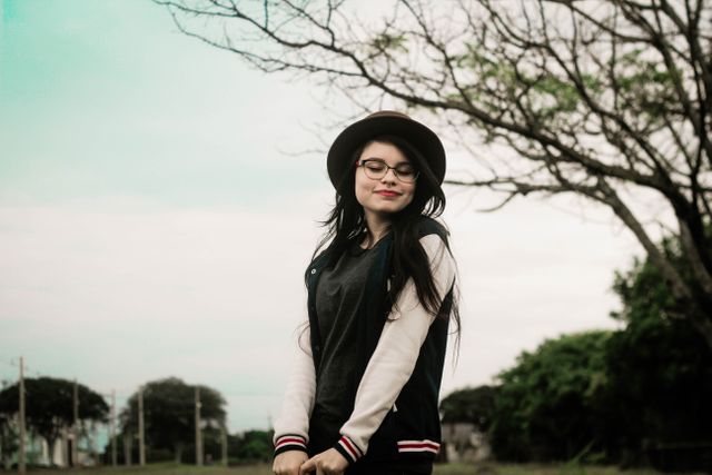 Young woman wearing hat and glasses stands outdoors with a serene expression, enjoying the natural surroundings. Ideal for use in lifestyle blogs, outdoor activity promotions, casual fashion advertisements, and articles about relaxation and self-care.