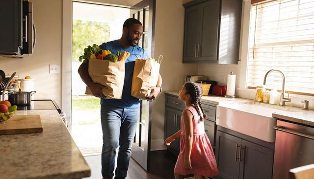 Smiling father carrying grocery bags into sunny kitchen greeting daughter