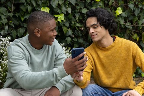 Diverse male friends sharing smartphone while sitting on bench by ivy hedge with white blooms