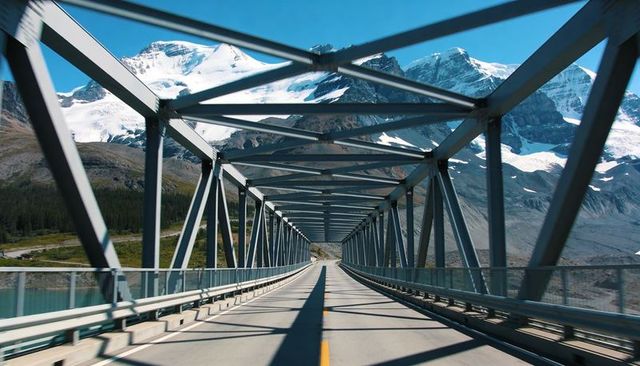 Driving across steel truss bridge toward snow-capped peaks and turquoise alpine lake