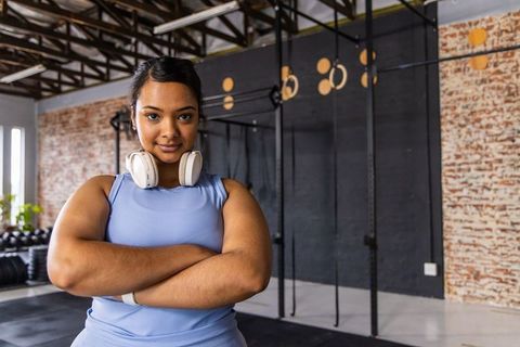 Confident asian woman standing arms crossed in gym wearing headphones and activewear