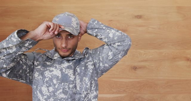 Soldier in camouflage uniform adjusting his cap against plain background, conveying readiness and focus. Suitable for use in military recruitment materials, articles on armed forces, websites dedicated to military service, and promotional content for military gear.