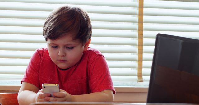 Young boy intensely focused on his smartphone while sitting at home with blinds in the background. Useful for topics related to children's technology use, screen time, digital communication, and home learning environments.