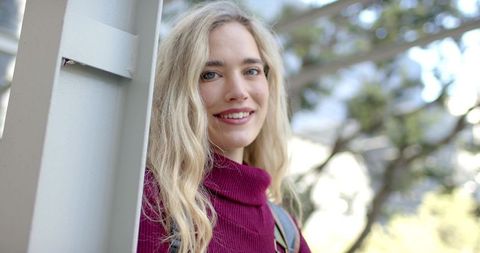 Young student leaning on post, smiling in burgundy turtleneck with backpack in park