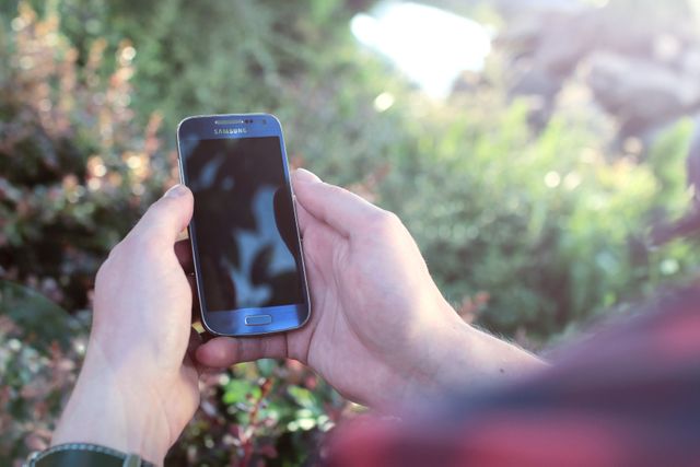 This image shows a person holding a smartphone outdoors with greenery in the background and sunlight filtering through the trees. Ideal for themes related to technology use in nature, outdoor connectivity, and modern communication. Perfect for blogs, advertisements, and articles focusing on mobile technology, apps, outdoor activities, and staying connected while on the go.