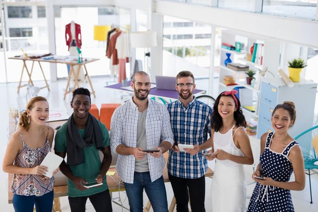 Group of young professionals standing together in a modern, creative office space. They are smiling and holding various items, suggesting a collaborative and friendly work environment. Ideal for use in articles or advertisements about teamwork, startup culture, modern office environments, and diversity in the workplace.