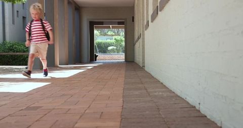 Schoolboy walking along sunlit brick corridor carrying backpack with copy space