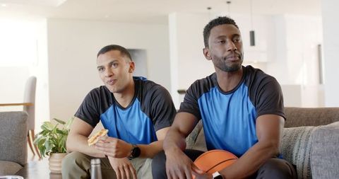 Diverse friends watching game on sofa holding pizza and basketball in modern living room
