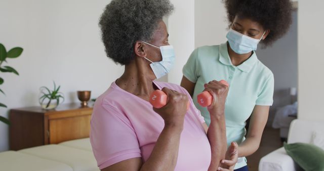 Senior woman lifting weights with assistance from caregiver at home while both wearing masks, focusing on fitness and health during COVID-19. Ideal for use in healthcare and fitness contexts, such as home care services, elderly exercise programs, and COVID-19 safety measures.