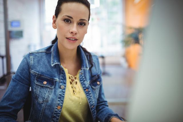 This image depicts a confident female executive sitting at her desk in a modern office environment. She is wearing a denim jacket and a yellow top, suggesting a casual yet professional look. This image can be used for business-related content, corporate websites, career blogs, and articles about women in leadership roles.