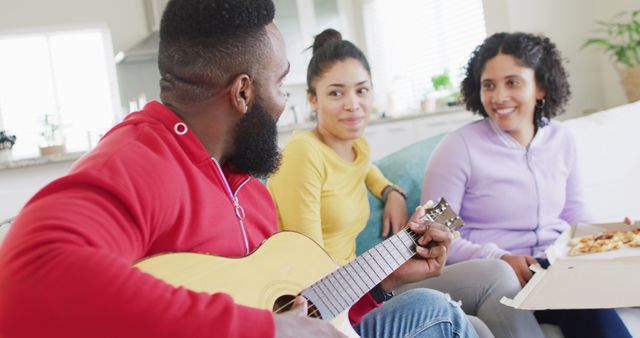 Young people relaxing and bonding in a casual home setting. One playing guitar while others enjoy pizza. Ideal for themes related to friendship, relaxation, leisure activities, and diverse companionship.