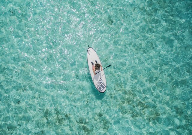 Top view showing person paddleboarding on crystal clear blue water. Useful for promoting travel destinations, active lifestyle blogs, water sport publications, outdoor adventure events, and summer vacation packages.