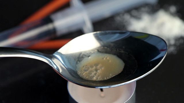 Video depicts heroin being prepared for use by heating in a silver spoon over a tea light candle. Syringe and white powder visible in the background suggest illicit drug use and addiction scene. Suitable for illustrating topics related to drug abuse, addiction, substance use, and its negative impacts.