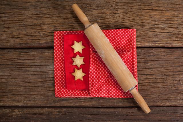 This image shows three star-shaped cookies placed on a red napkin with a wooden rolling pin beside them on a rustic wooden table. Ideal for holiday baking blogs, Christmas recipe websites, festive greeting cards, and culinary magazines.