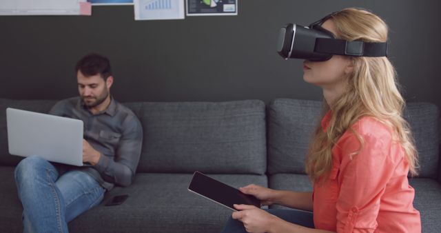 Woman sitting on sofa using VR headset, engaged in immersive experience. Man working on laptop in background, demonstrates multitasking and modern technology in creative workspace. Perfect for tech-oriented and office environment concepts.