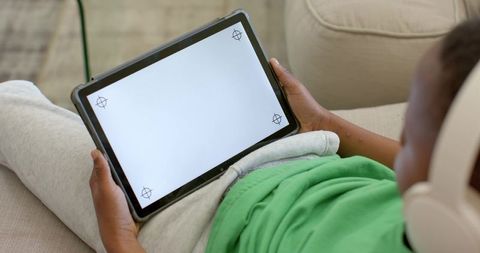 Child reclining on couch holding tablet and wearing headphones for online learning at home