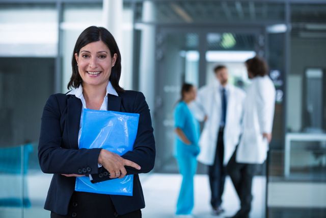 Businesswoman standing in hospital premises holding a file, smiling confidently. Medical staff in background, suggesting collaboration between business and healthcare. Ideal for use in articles about healthcare management, corporate roles in medical settings, and professional success in the healthcare industry.