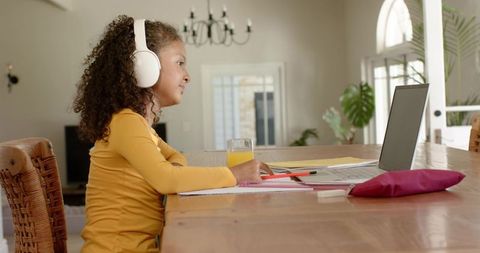Girl studying at home with headphones and laptop on wooden table in sunlit dining room