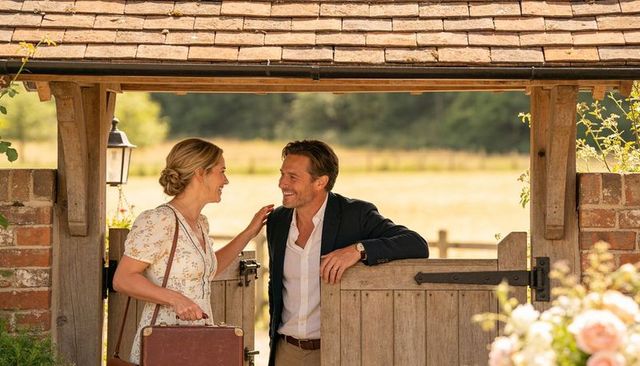 Smiling couple at rustic cottage gate holding vintage suitcase, romantic summer countryside moment
