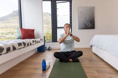 Mature man meditating on yoga mat in sunlit minimalist bedroom for wellness