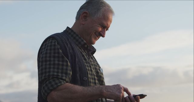 Elderly man smiling while using his smartphone outdoors against a pastel-colored sky at sunset. Suitable for use in contexts of technology adoption among seniors, promoting digital literacy, outdoor leisure, or showcasing positive aging and connection. Ideal for advertisements, brochures, or websites targeting seniors and modern technology use.