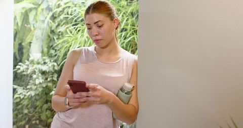 Leaning woman checking phone after workout holding bottle wearing fitness tracker