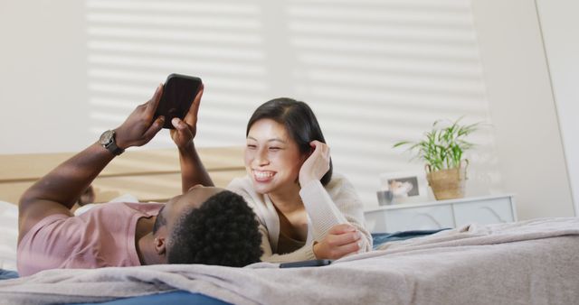 Happy diverse couple using smartphone and lying in bedroom. Spending quality time at home concept.