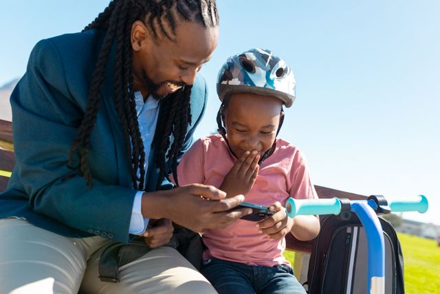 Happy african american single father sharing smartphone with son wearing protective helmet. unaltered, telecommunications, enjoyment, family, lifestyle and togetherness concept.
