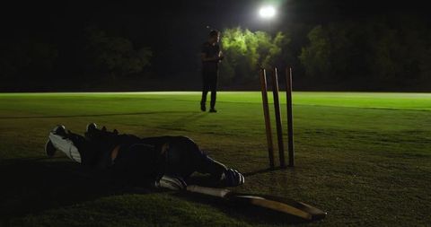 Lying batsman face-down on night cricket pitch under floodlights with bat and stumps