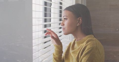 Peering through blinds mid-adult woman gazing out window in mustard sweater, contemplative