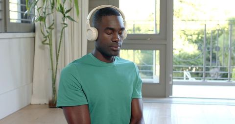 African American man meditating with headphones by balcony doors in bright modern home