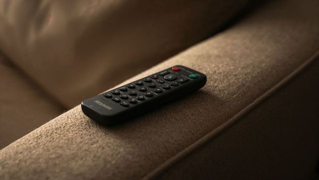 Black tv remote resting on brown sofa armrest with warm lighting and visible buttons