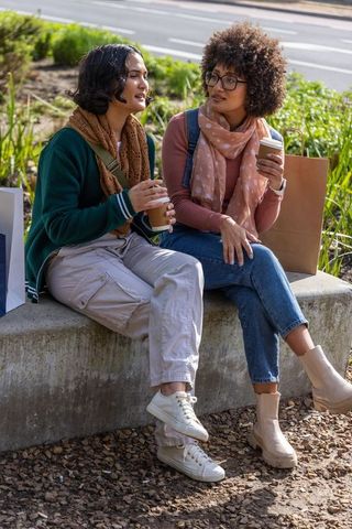 Two friends chatting over coffee on urban ledge with shopping bags and casual fall style