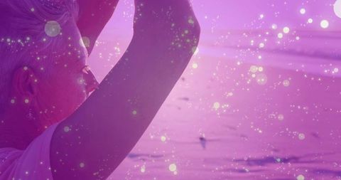 Senior woman raising arms toward ocean horizon at sunset with magenta sparkling bokeh