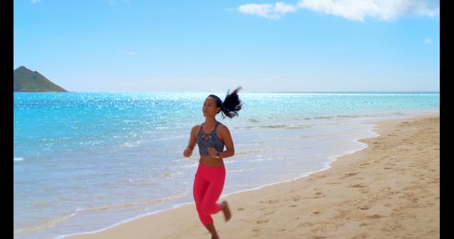 Young biracial woman jogging on a sunny beach, with copy space. Her active lifestyle is evident as she enjoys a run by the sea.