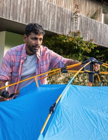Indian man assembling blue camping tent in backyard adjusting yellow poles and hub