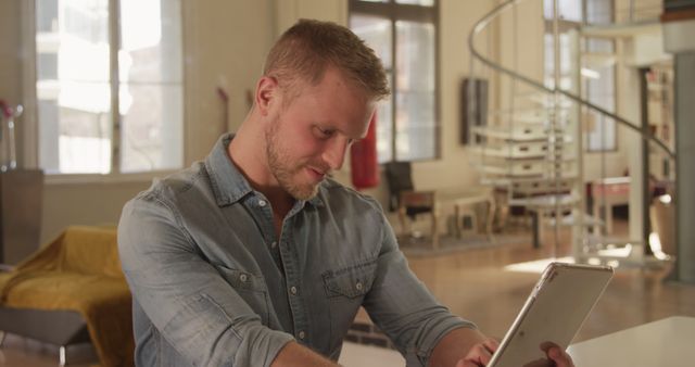 Man engaging with digital tablet device in a contemporary home interior wearing casual denim shirt. This visual illustrates modern technology usage in a residential setting. Suitable for lifestyle blogs, technology advertisement, and articles on home office productivity.