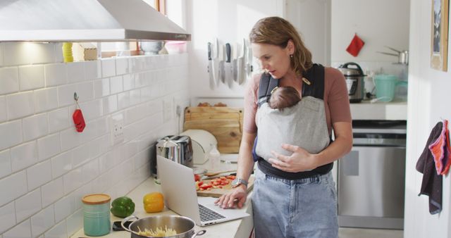 Mother is carrying her baby in a baby carrier while working on a laptop and cooking in the kitchen. The scene illustrates the balance of work and family life, capturing modern parenthood. Useful for themes related to work from home, parenting tips, multitasking, and family life dynamics.