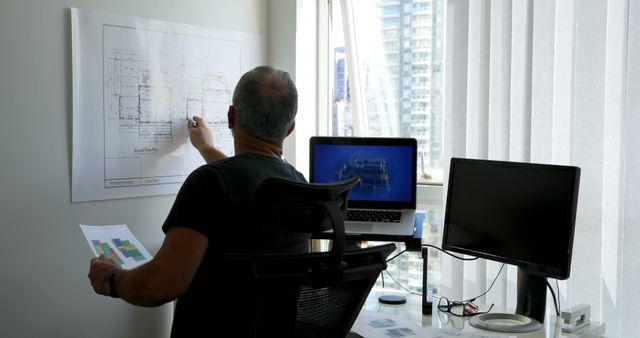 Architect is analyzing building plans while referencing a computer screen in a sleek, modern office. This stock photo can be used for promotional materials, articles on architecture and design, urban development presentations, or website images for construction and architectural firms.