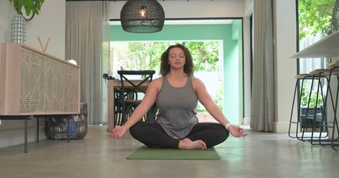 African American woman meditating on green yoga mat in modern living room wearing smartwatch