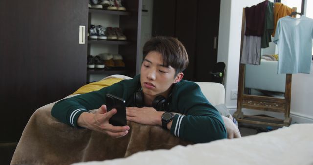 Young man in casual attire relaxing on the sofa while checking his smartphone. Headphones around his neck indicating he might be listening to music or podcasts. Room features modern home interior with organized closet and hanging clothes. Useful for lifestyle, technology, and casual home settings content.