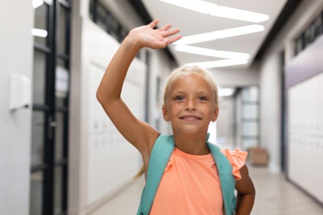 Smiling caucasian elementary schoolgirl waving hand while standing in school corridor. unaltered, education, childhood, happiness, waving, greeting and school concept.