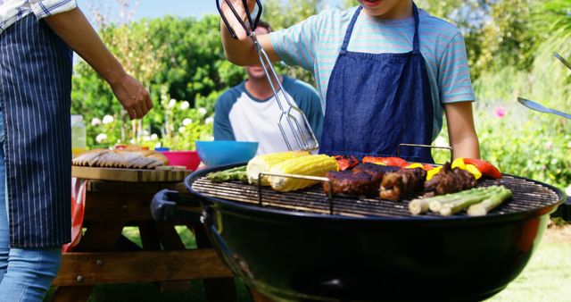 Children wearing aprons grilling vegetables and meat on a barbecue grill in a green backyard. Family members enjoying picnic table with food in sunny weather. Ideal for family, outdoor, and summer themed promotions, blogs, and advertisements focusing on bonding, outdoor activities, and grilled food.