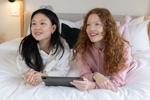 Two friends relaxing on bed sharing tablet, smiling and browsing in cozy loungewear